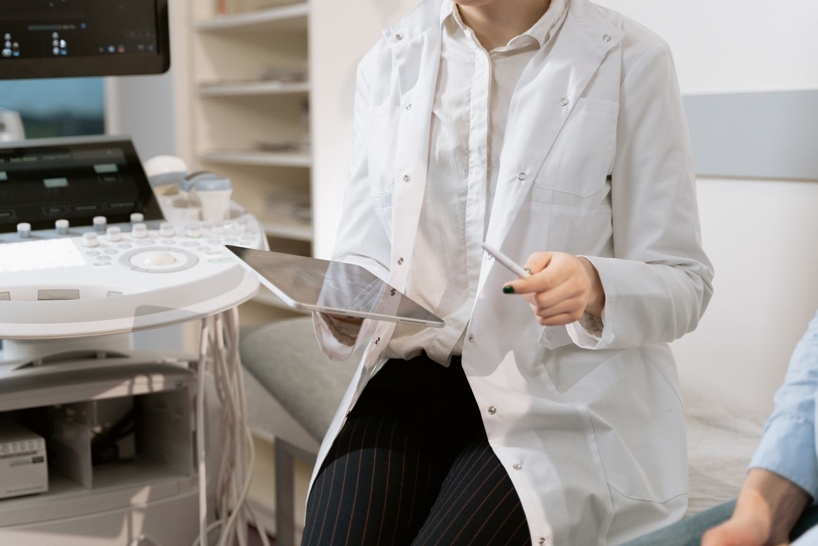 Sala de exame com aparelho de ultrassom e médica de jaleco branco segurando um tablet